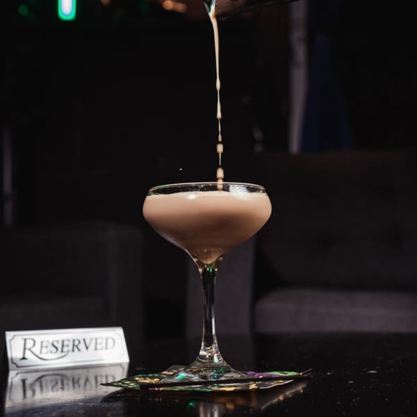 A creamy brown cocktail being poured into a coupe glass on a dark table with a "Reserved" sign.