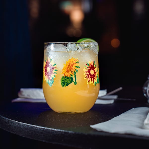 A vibrant yellow cocktail in a sunflower-decorated glass with a salted rim and lime wedge.