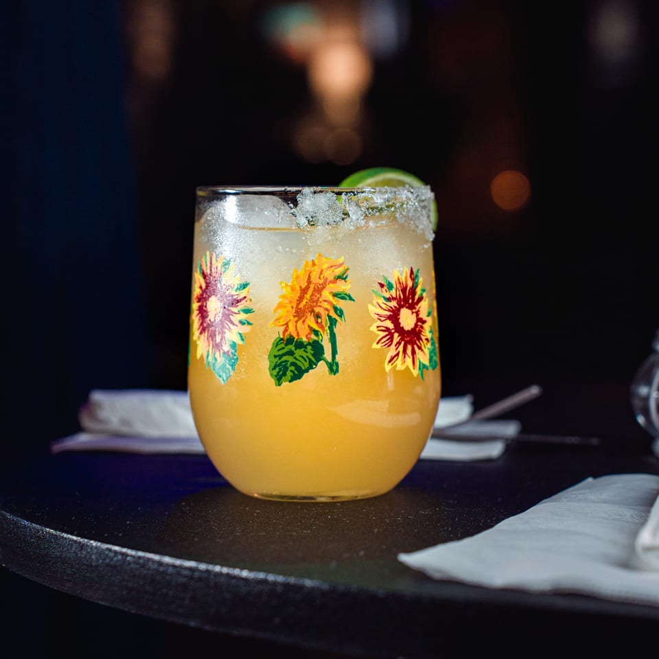 A vibrant yellow cocktail in a sunflower-decorated glass with a salted rim and lime wedge.