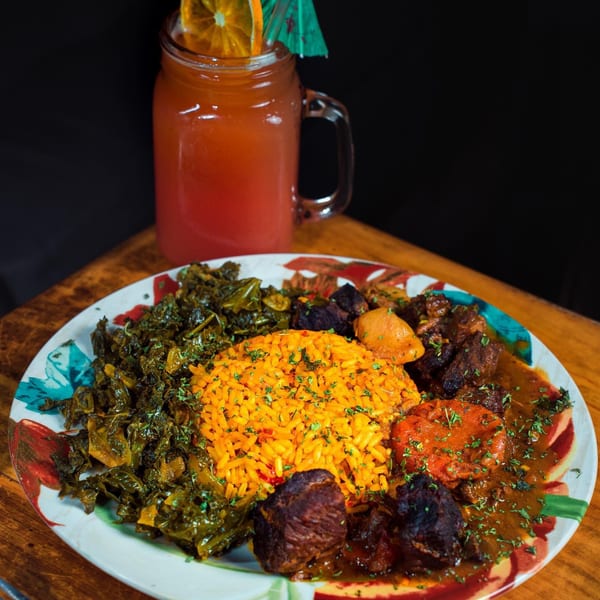 African dish with yellow rice, stewed greens, meat, and a drink with an orange slice.