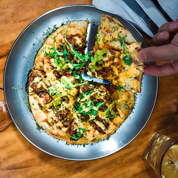 A hand takes a slice of African-style pizza topped with meat, cheese, and arugula from a metal plate.