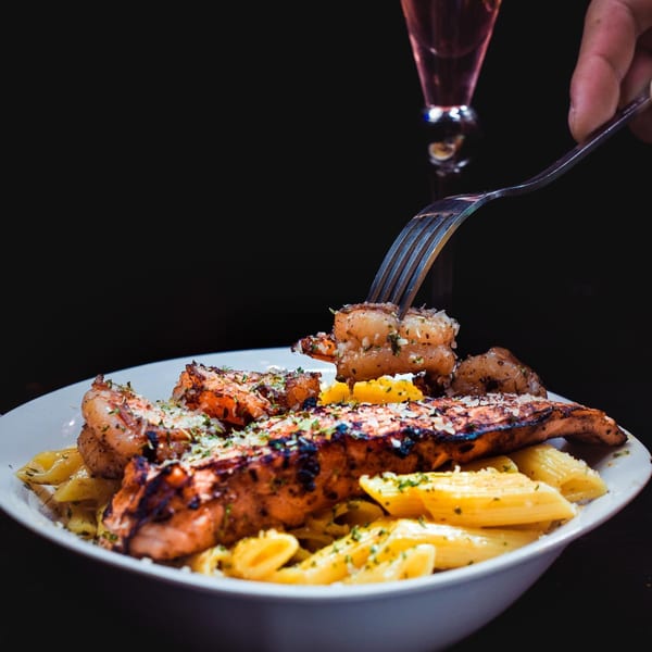 Grilled salmon and shrimp pasta dish with a fork lifting a shrimp, and a wine glass.