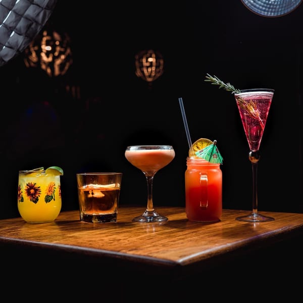 Five colorful cocktails, including a red drink with a paper umbrella, on a dark wooden bar.