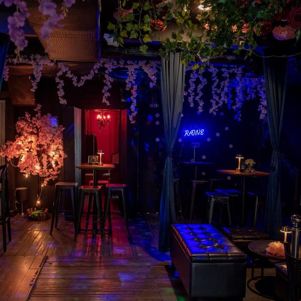 A dimly lit, vibrant bar interior with neon signs, floral decor, high-top tables, and stools.