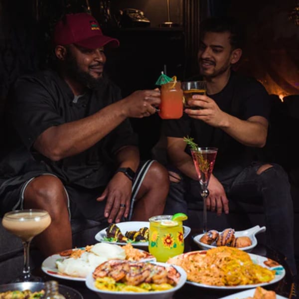 Two men toasting drinks over a table laden with various African dishes and cocktails.