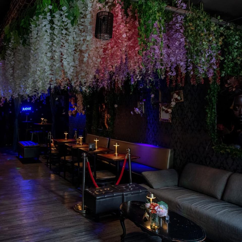 Dark, moody restaurant interior with hanging floral decor, booth seating, and glowing table lamps.