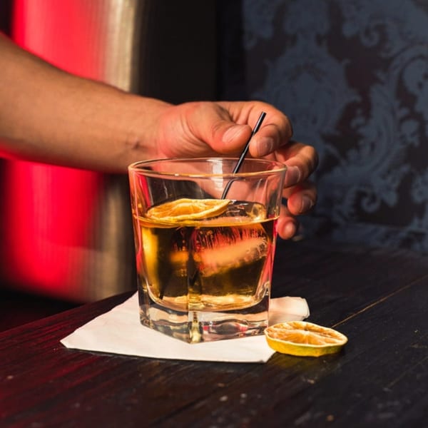 A hand stirs a cocktail with dried orange slices and a large ice cube on a dark bar.