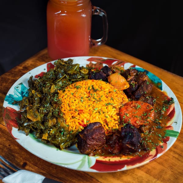 African dish with orange rice, stewed greens, meat, and vegetables on a colorful plate with a drink.