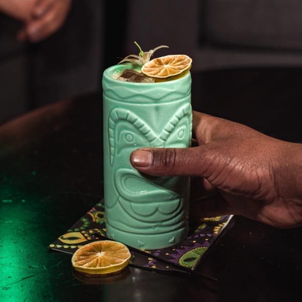 A hand holds a teal tiki mug with a drink garnished with dried citrus and a plant.