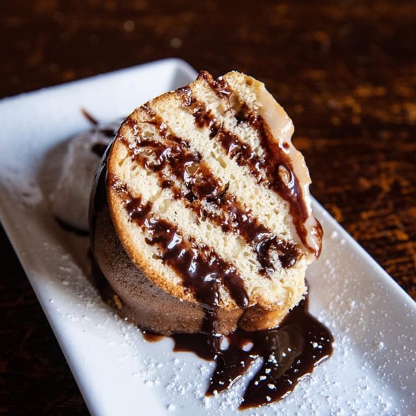 A slice of bundt cake with chocolate drizzle and powdered sugar on a white plate.