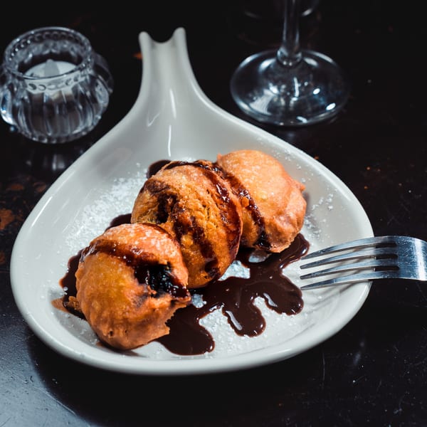 Three African fried dough balls drizzled with chocolate sauce and powdered sugar on a white plate.