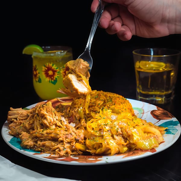 A hand holds a fork with chicken over a plate of African food, with drinks in the background.
