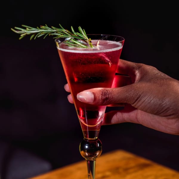 A hand holding a tall, elegant glass with a red cocktail garnished with a sprig of rosemary.