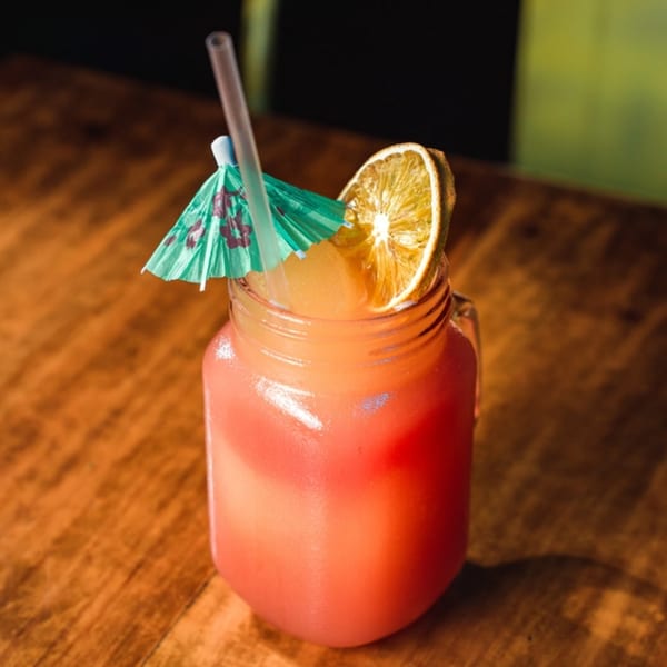 A vibrant red and orange cocktail in a mason jar, garnished with a dried orange slice and a paper umbrella.